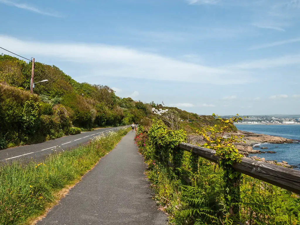 A pavement path, with the road to the left and a wooden fence on the right, near the end of the walk from Penzance to Mousehole. There is tall grass between the road and the path. The sea is visible below to the right. 