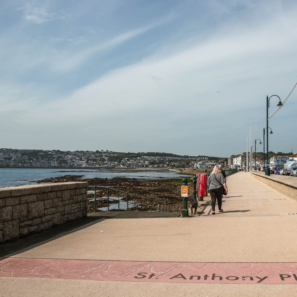 The Strat of the promenade in Penzance, with the sea wall to the left. There is a woman walking on the promenade.