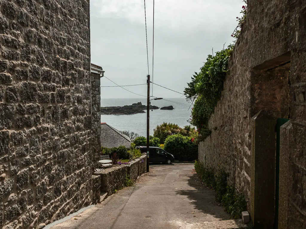 A road leading downhill in between the stone walled buildings, with the sea ahead. 