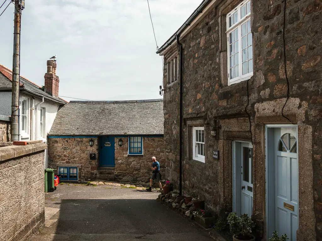 A back street, with a stone cottage ahead, and stone walled house to the right. There is a man walking in between the buildings. 