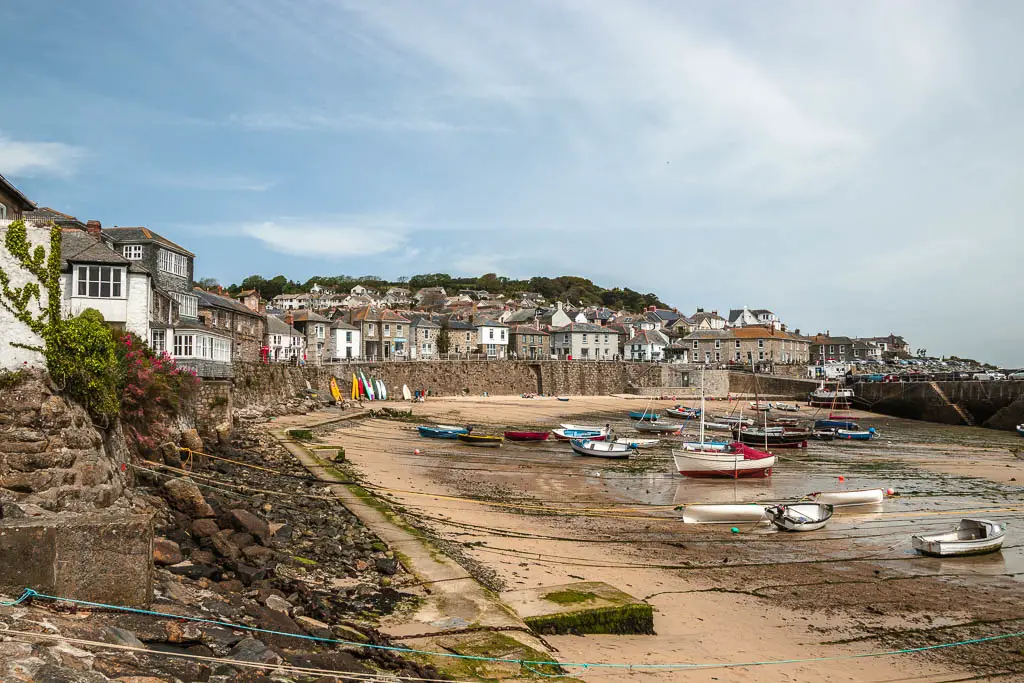 Looking across the bay of mousehole at low tide. There are lots of boats resting on the sea bed. There is a stone wall on the left of the bay. There are lots of houses behind the wall.