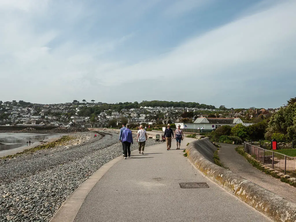 People walking along the path as it curves around the shingle beach on the left. There are houses in the distance up a small hill.