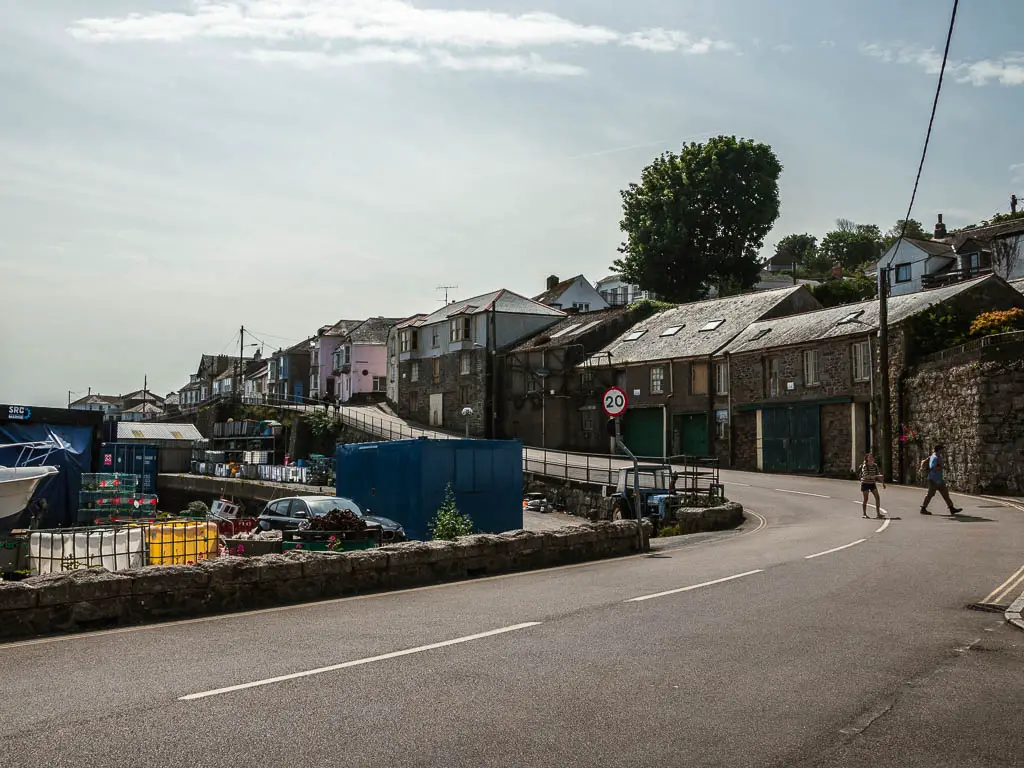 A road as it curves ahead and uphill to the left. There are a couple of people walking on the road. The road is lined with houses on the right side. 