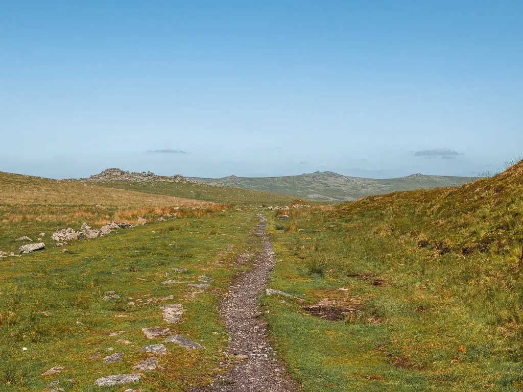 A very narrow rocky path running through the grass with a view to King's Tor in the distance, on the walk along the Princetown Railway.