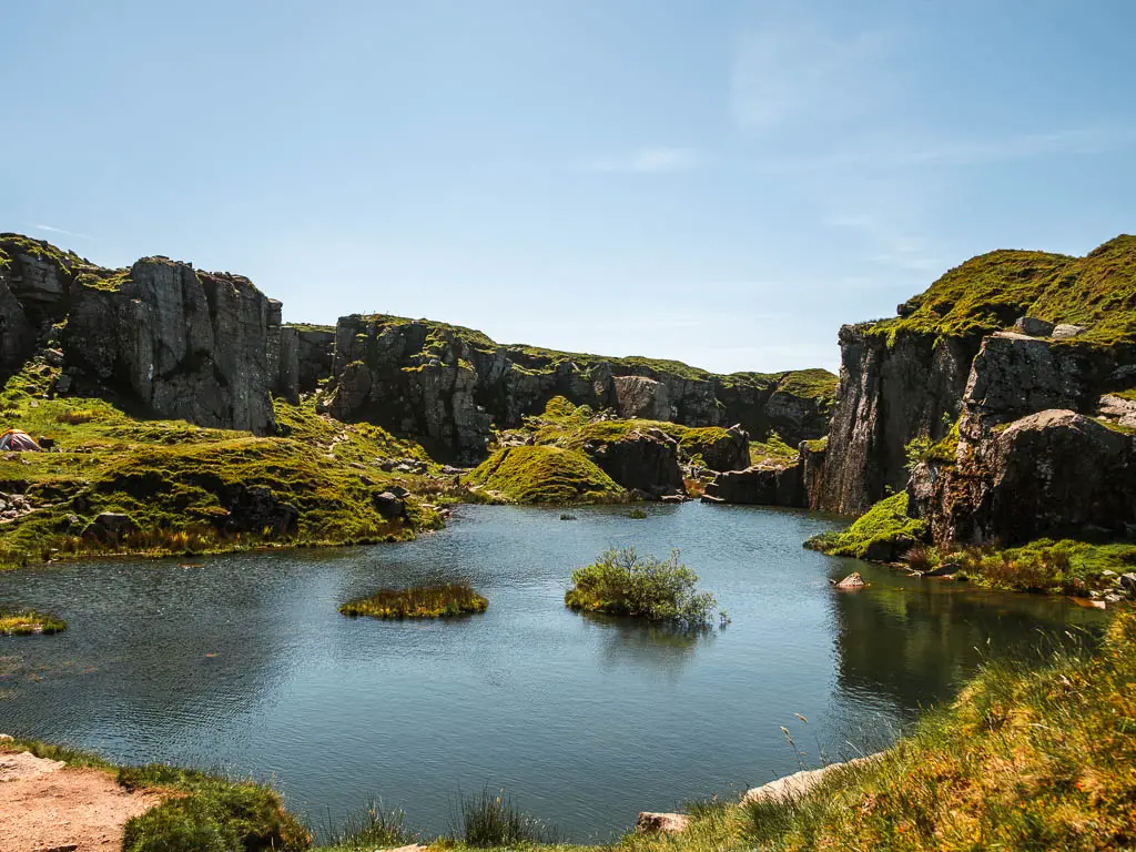 Looking across the calm water of Foggintor Quarry, surround by rock cliff, covered in grass, on the circular railway path walk from Princetown