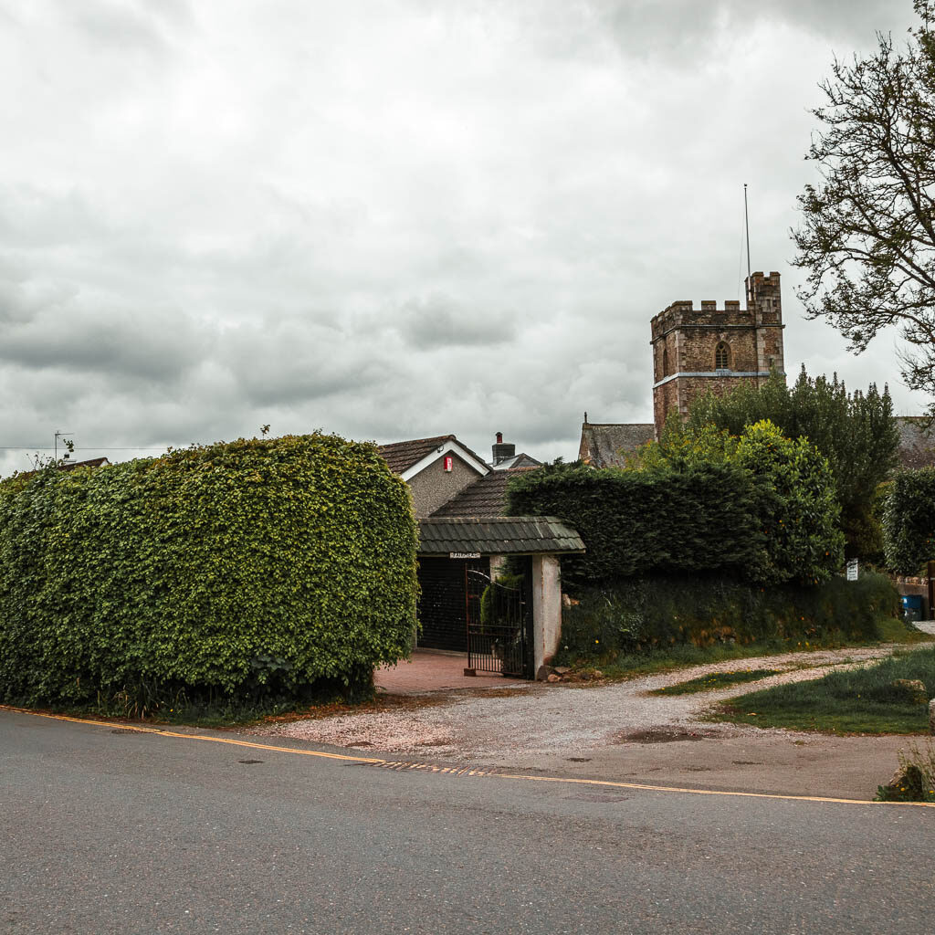 Looking across the road to a hedge and part of a church visible coming up from behind it.