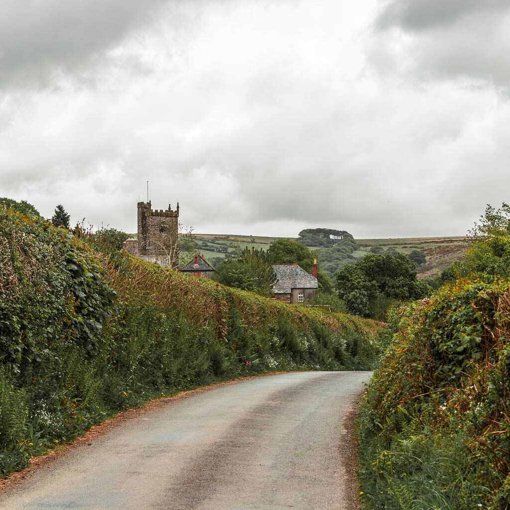 A country road as it curves to the right, lined with hedges and part of a church visible ahead to the left. 