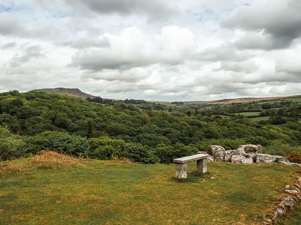 A stone bench on the flat green hill top, with a view over the trees to Sheepstor, and the end of the circular walk.