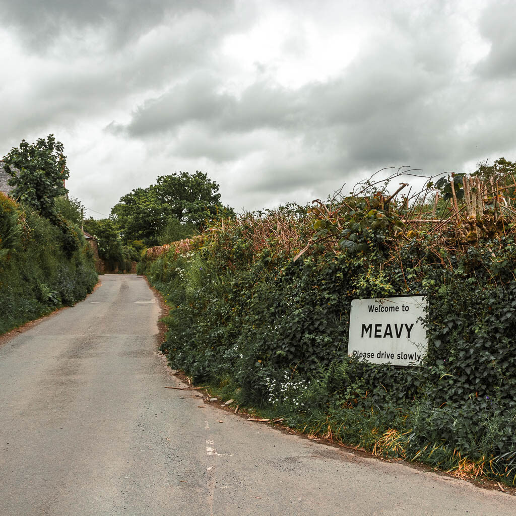 A country road lined with hedges and a white sun on the right which says 'Welcome to Meavy'.