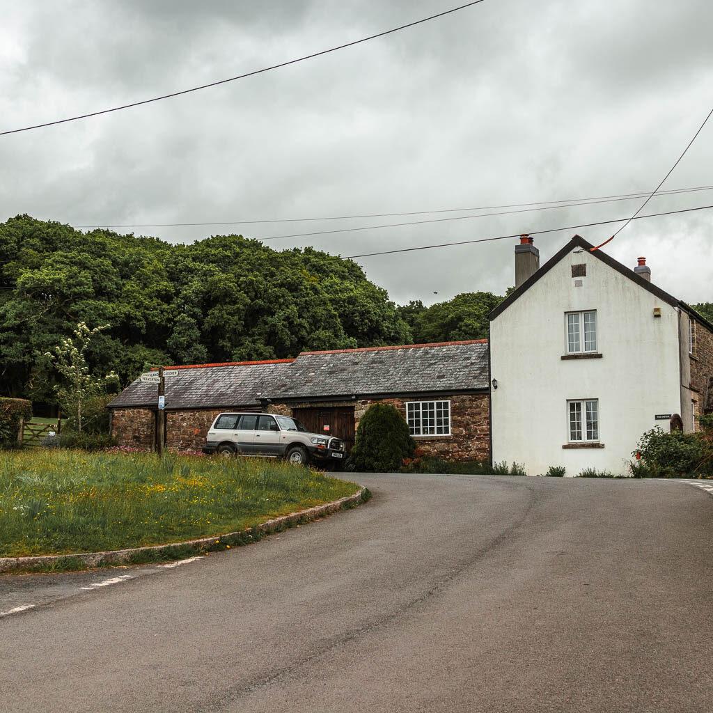 A road, with a green island on the left and a house ahead. The house has a white wall, with a stone walled extension. There is a car parked out side the house.