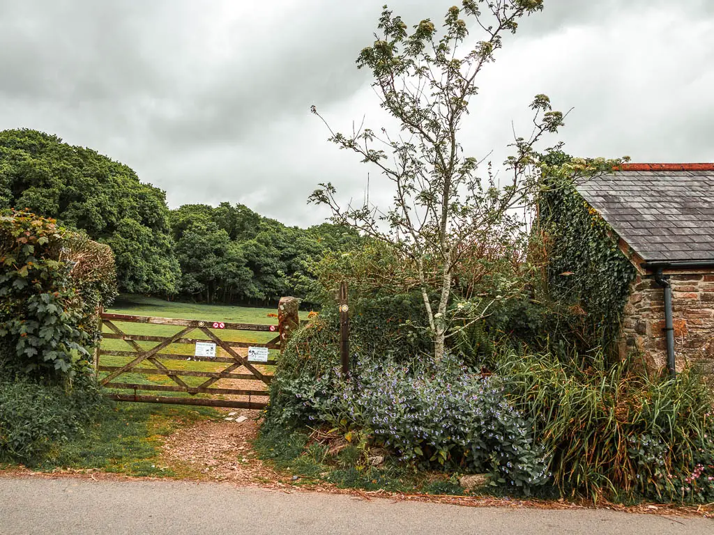 A wooden gate on the left, with bushes and hedge on either side, and part of a stone building on the right.