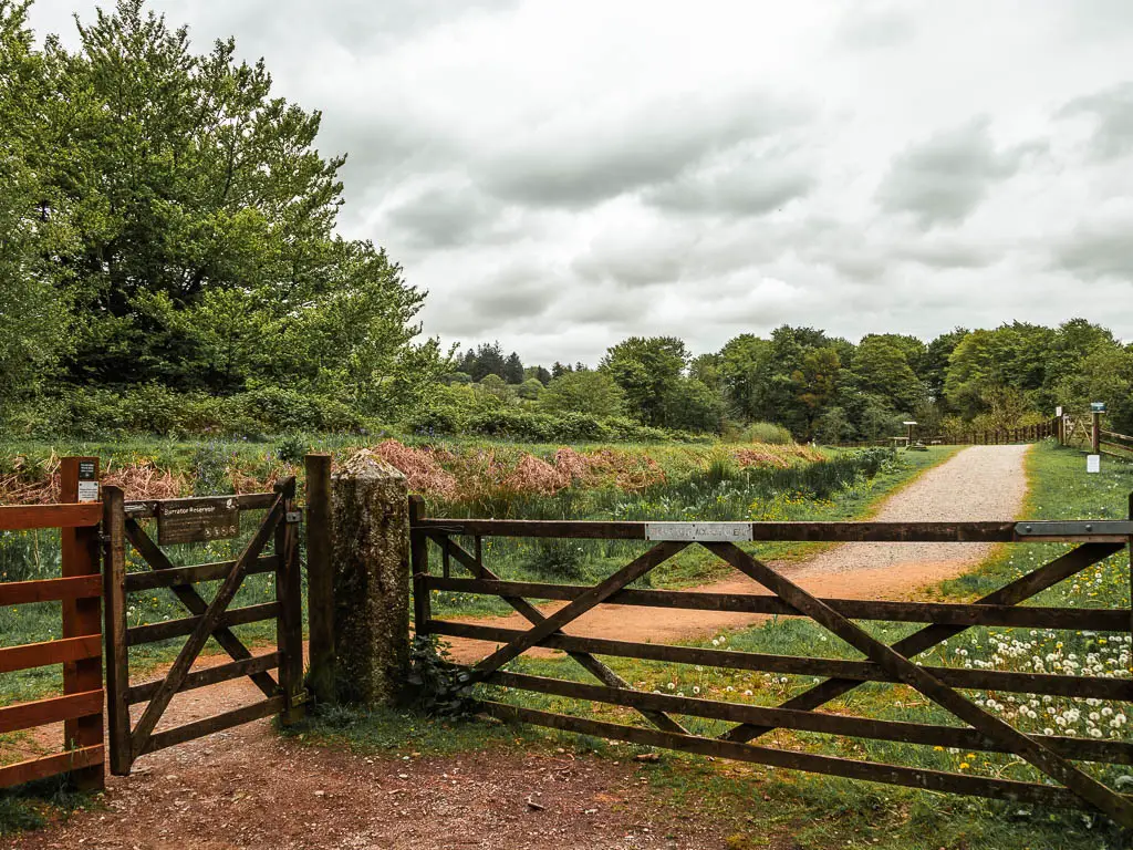 A wooden gate leading to a wider path, and some trees further ahead on the path.