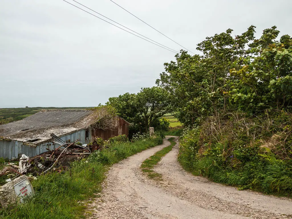 A gravel type road, winding ahead, with an abandoned shed on the left and bushes and trees to the right. 