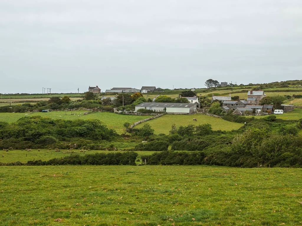 Looking down along the large grass field, to a hedge on the other side, then a few more field beyond that leading to a cluster of houses.