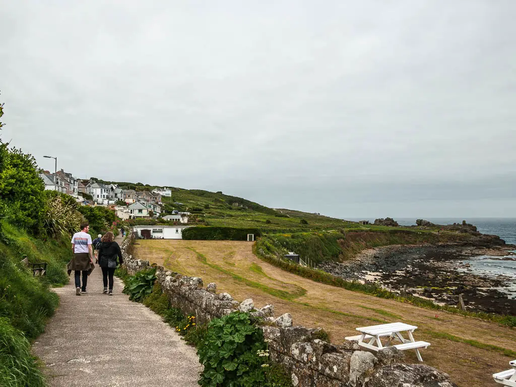 A path on the left, with a stone wall to the right, and a large green to the right of that. There are a couple of people walking on the path. There are white picnic benches on the green.