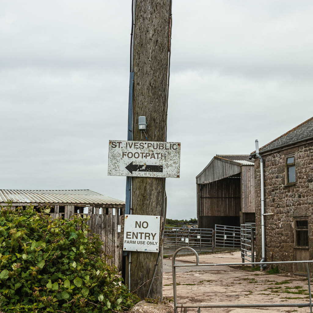 A sign pointing to the St Ives footpath on the circular walk back from Zennor. The sign is on a wooden pole in front of some farm buildings.