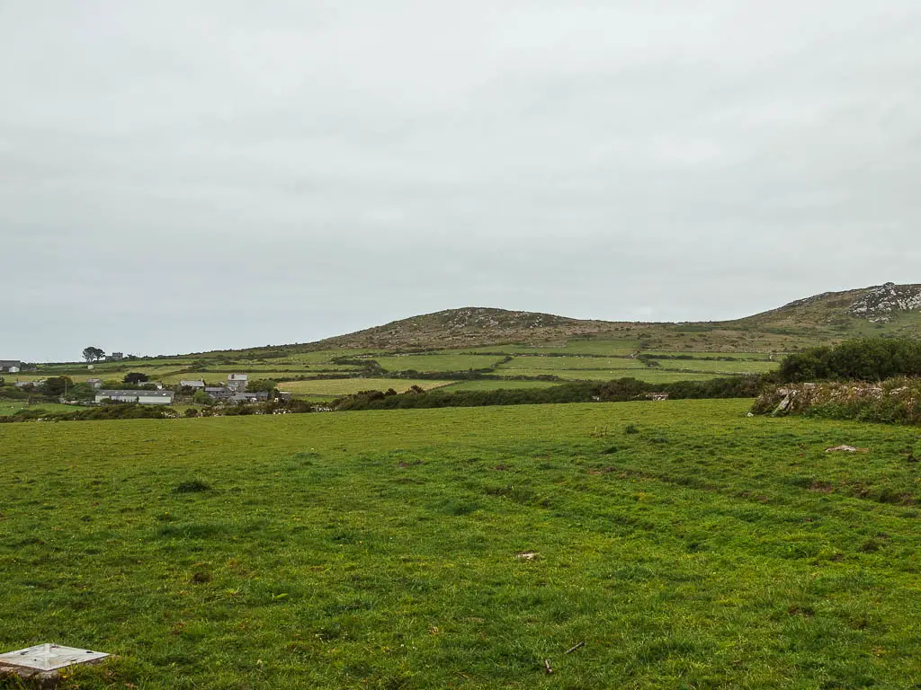 A large grass field, with a hill ahead in the distance and a small village visible to the left in the distance. 