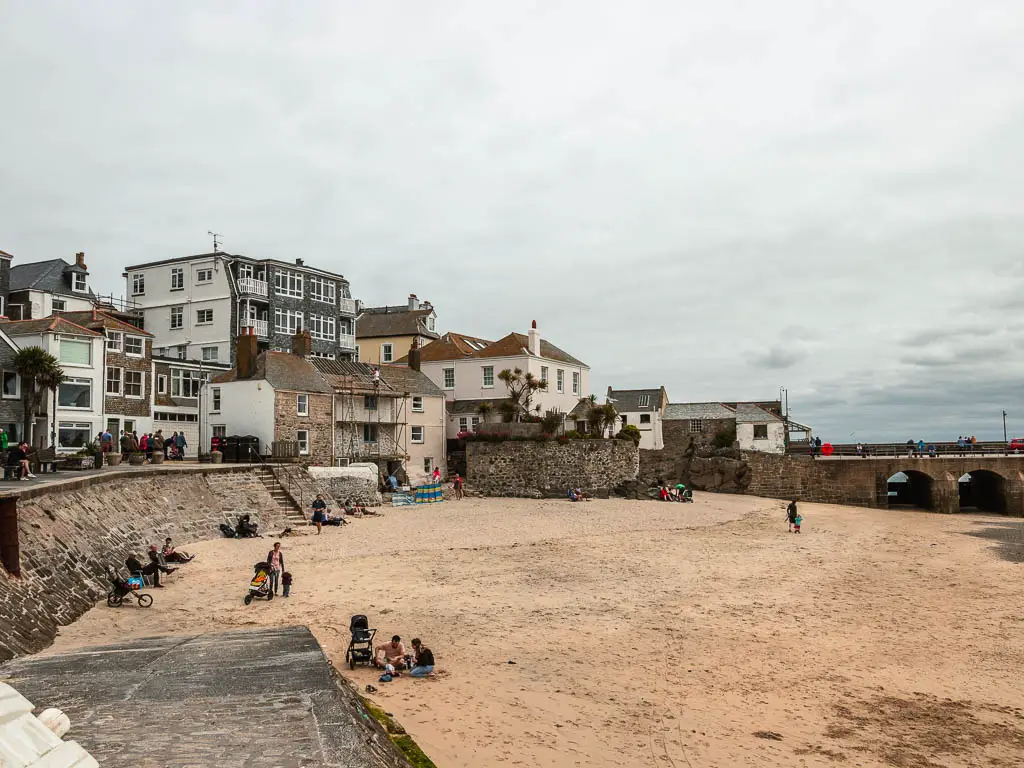 A sandy beach cove in St Ives at the start of the walk to Zennor along the coast path. The beach is enclosed by a stone wall on the left, with the road above it. The road is lined with buildings. There are a few people sitting on the sand.