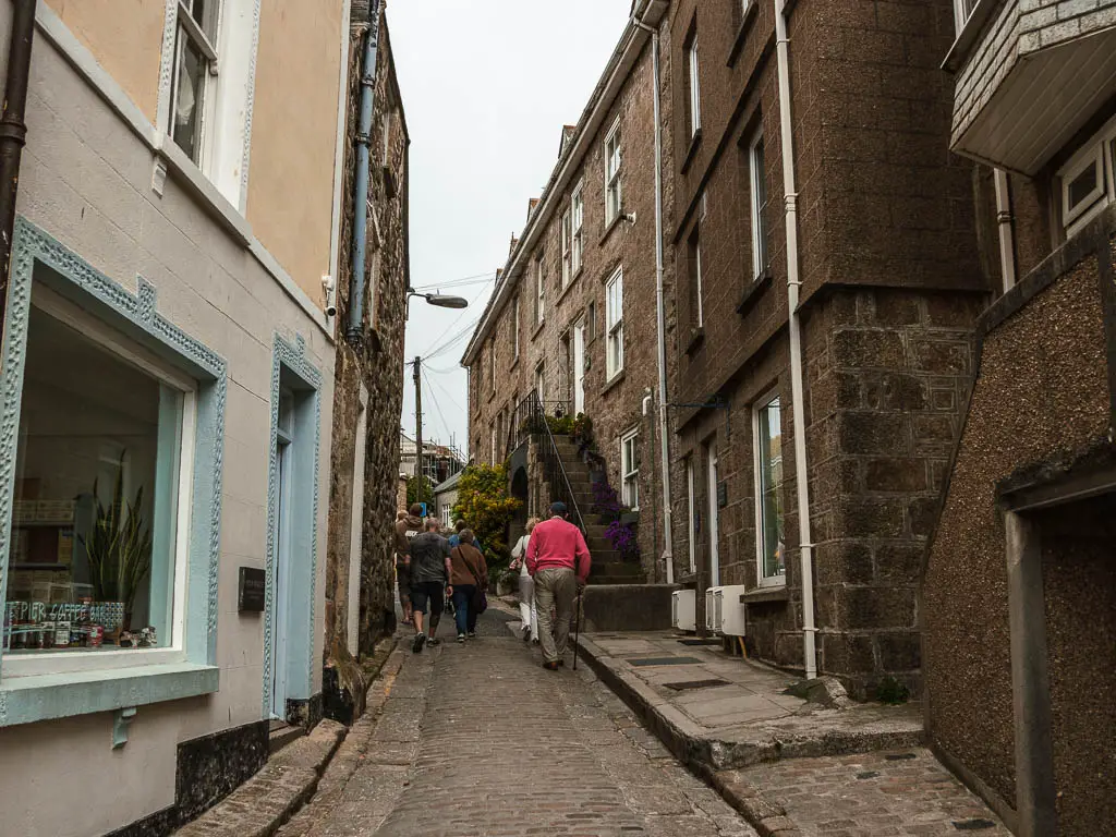 A narrow alley leading uphill, lined with shops and houses. There are people ahead walking up the hill.