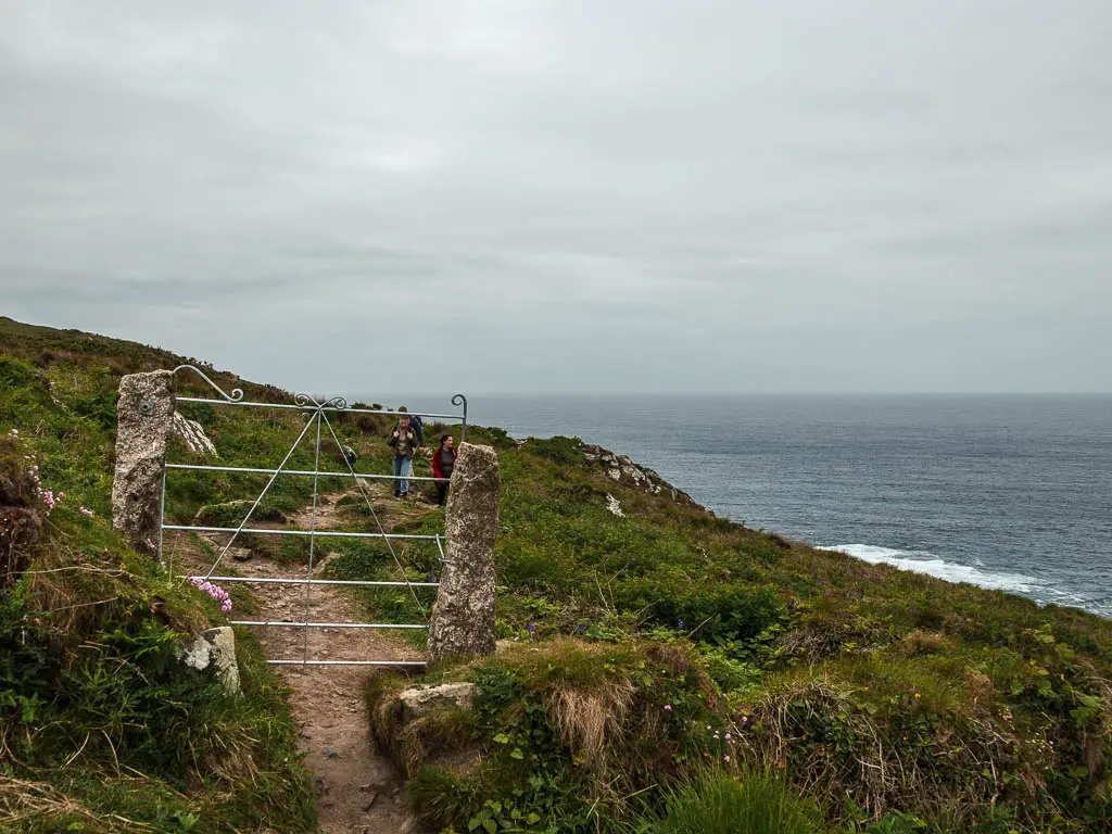 An ornate metal gate in the middle of the trail, surround by grass and greenery along the St Ives to Zennor walk. The sea is to the right, and there are some people walking on the trail on the other side of the gate. 