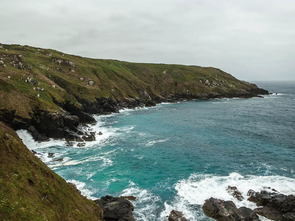 A rugged cliff cove on the coastal walk from St Ives to Zennor. The cliffs are covered in grass as they slope down, then become black rocks as they meet the rough sea. 