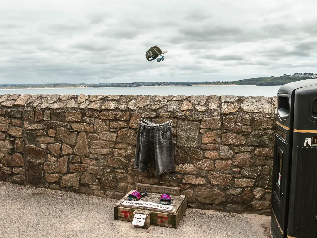 A stone wall with an optical illusion invisible man in front of it, at the start to the walk from St Ives to Zennor. There are shorts, then a hat and sunglasses floating in the air. 