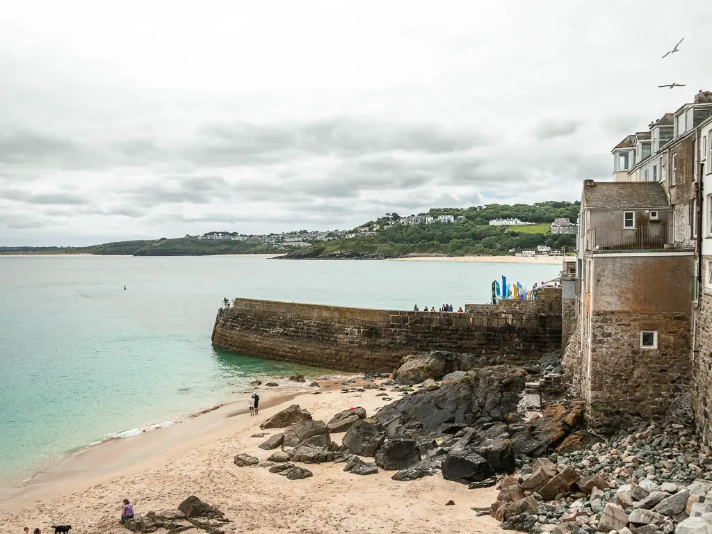 Looking down to the sandy beach cove in St Ives at the start of the walk to Zennor along the coast path. The beac h has lots of rocks, and is secluded off with a wall and stone houses. The sea is pale turquoise in colour. 