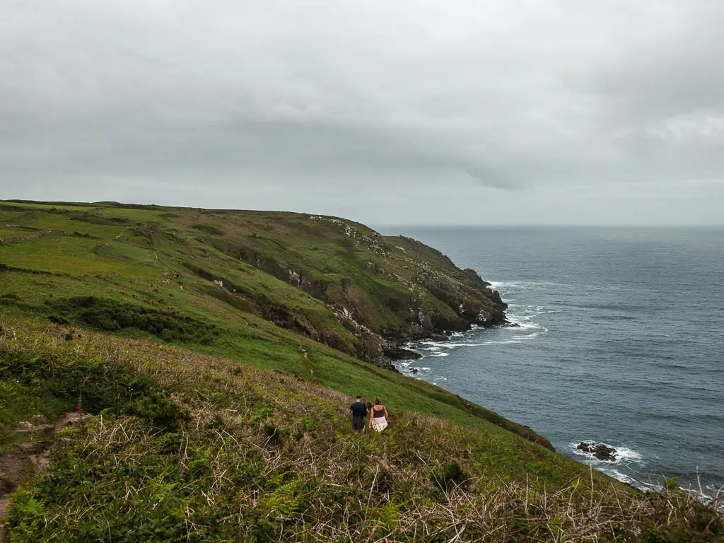 Looking down and along the undulating grass covered clifftop on the coastal walk from St Ives to Zennor. The cliffs slope down to meet the blue sea on the right. There are a couple of people walking on the trail running along the top of the cliff.