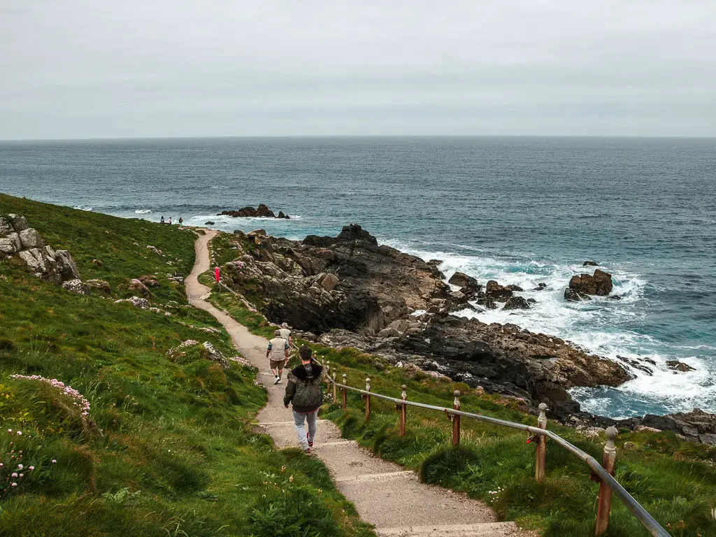 Steps leading downhill, with black rocks and sea to the right, and grass to the left. There is a railing on the right side of the steps and people walking down them.