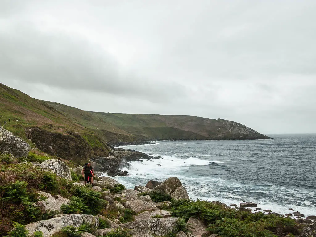 At the bottom of the cliffs, with lots of rocks forming the path ahead, and the cliff cove curving around from left to right, on the St Ives to Zennor coastal walk. There are a couple of people ahead, walking across the rocks. 