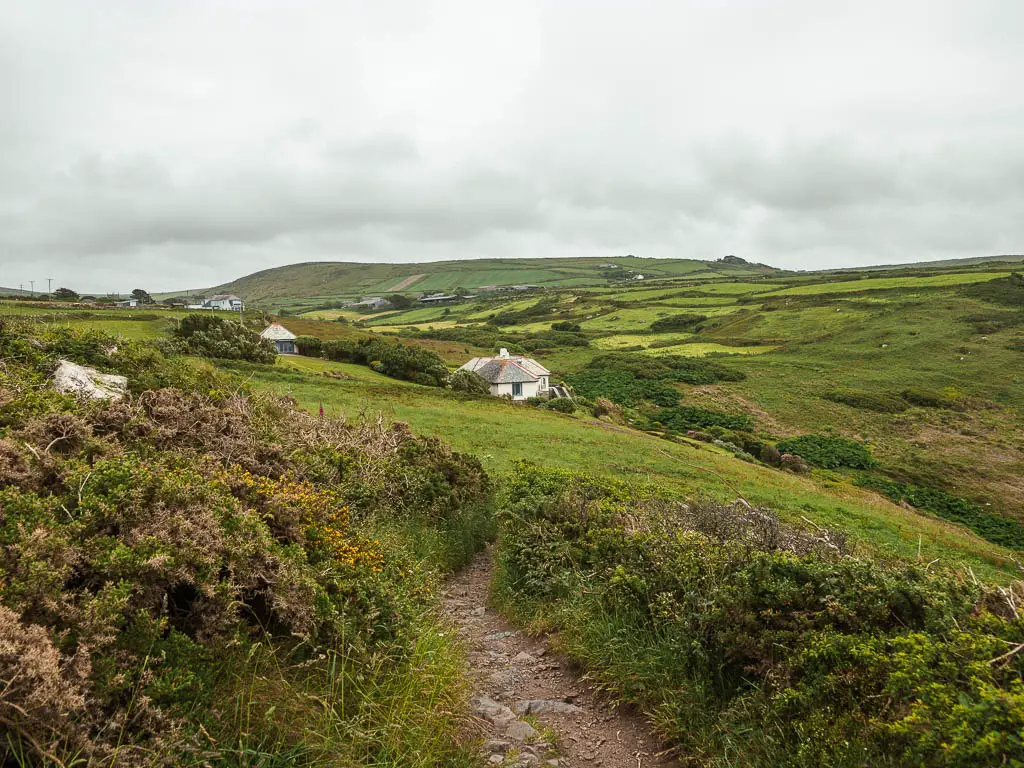 A small dirt trail soured by grass and green plants, with a small white cottage ahead and hills in the distance. 