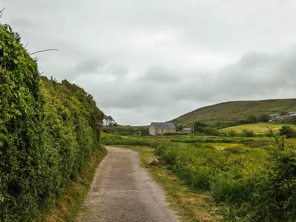 A small road path, with a tall hedge on the left and field leading to hills on the right. There is a small cottage ahead in the field. 