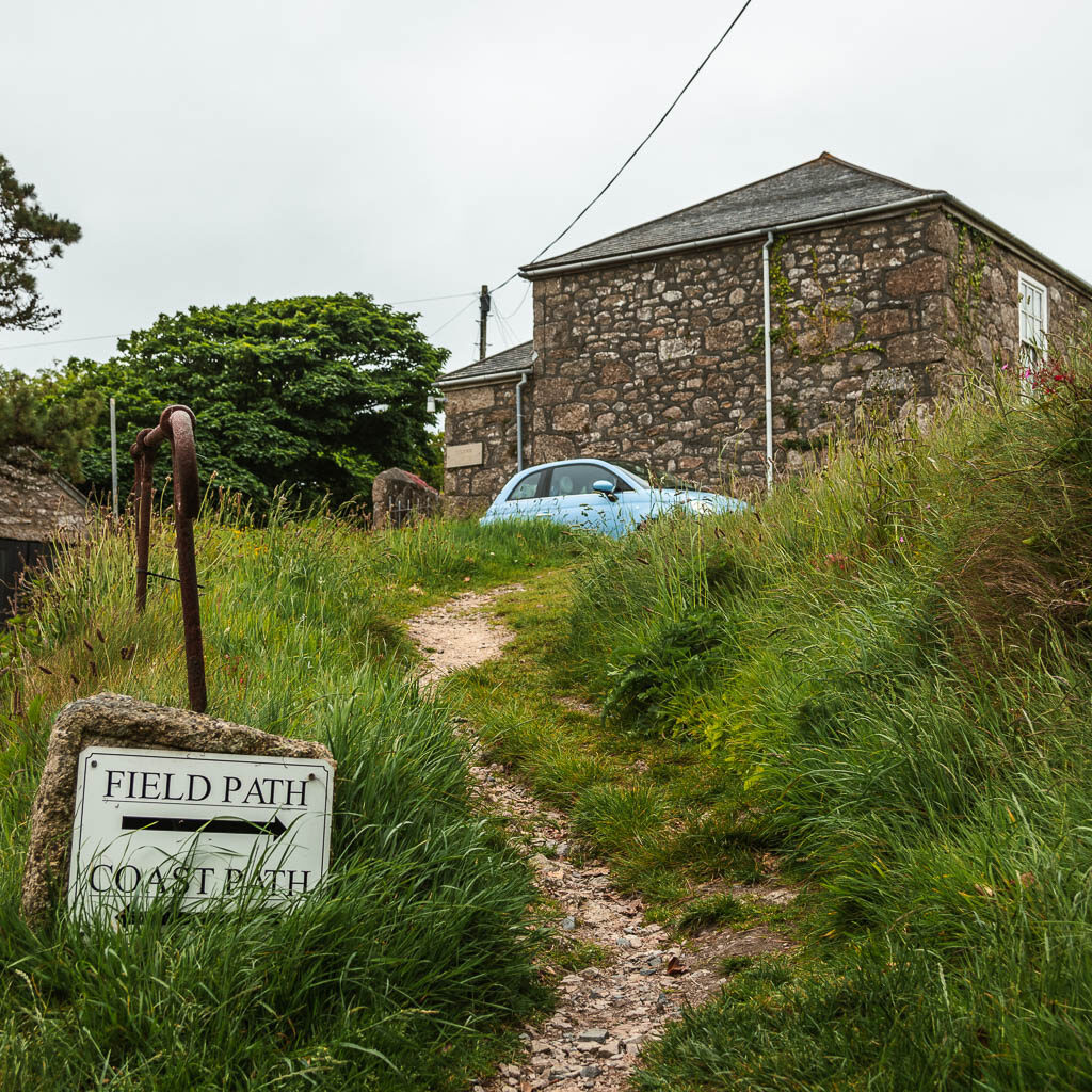 A narrow trail leading up through the tall grass, with a stone building and light blue car ahead. There is a sign next to the trail pointing to the field path. 