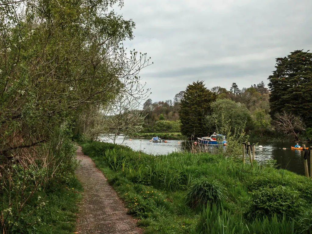 A path on the left, with a grass bank and river to the right. A few boats are on the river.