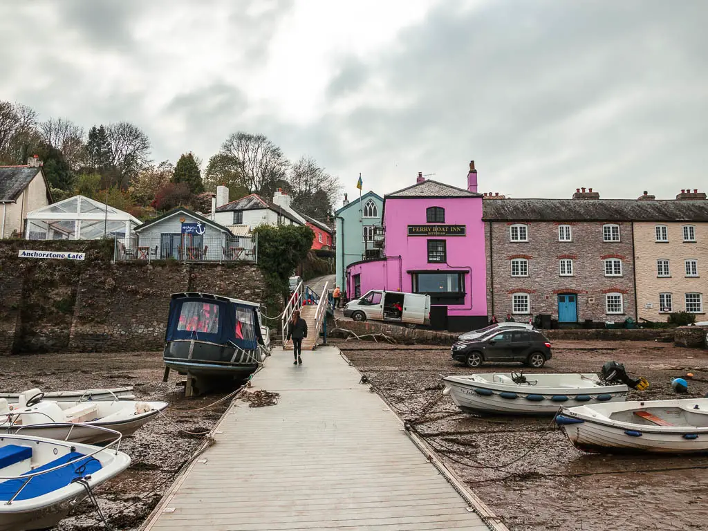 Looking along the pier to Dittisham, with the pink façade of the Ferry Boat Inn pub. There is a person walking along the pier, which is also lined with lots of small boats.