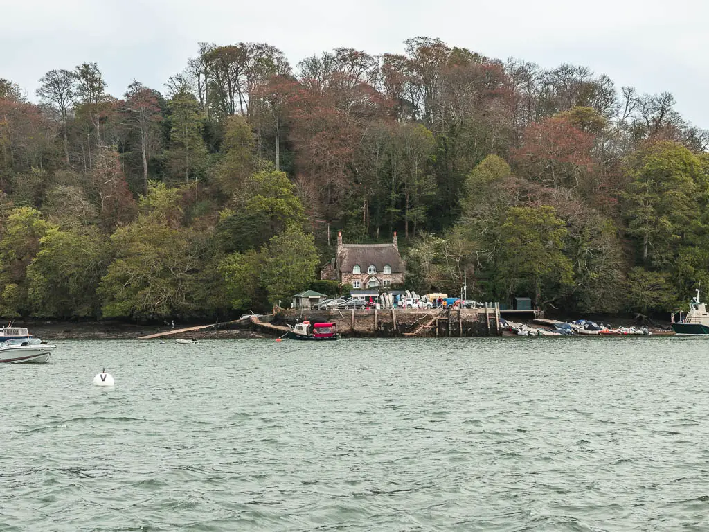 Looking across the choppy river to a thatched roofed cottage on the other side at the end of the Totnes to Dittisham walk. There is a platform and pier in front of the cottage, and lots of trees behind.