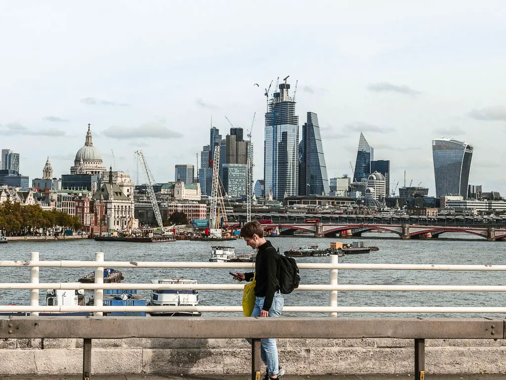 A view past the bridge railings and along the river, to the city of London skyline with all the tall skyscrapers. There is a person walking along the bridge.