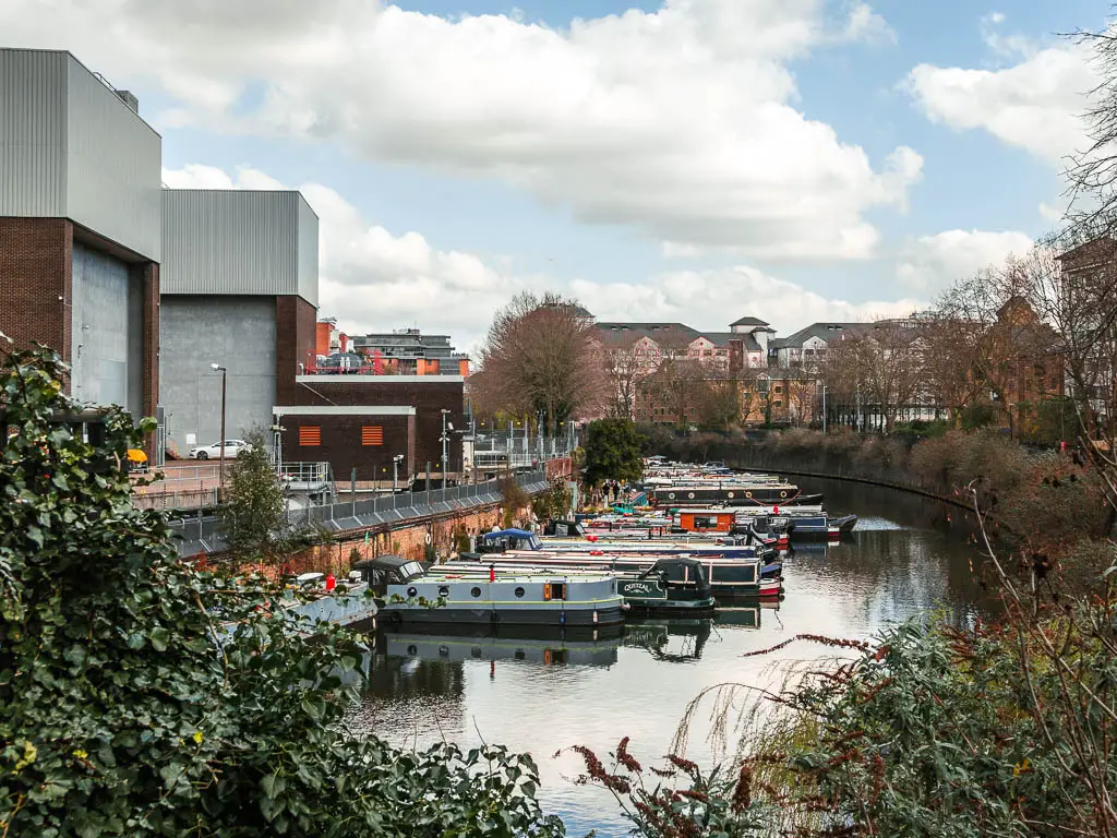 Looking over the bushes to the regents canal, with factory buildings on the left of the path, and lots of barges lined up, partway through the walk.