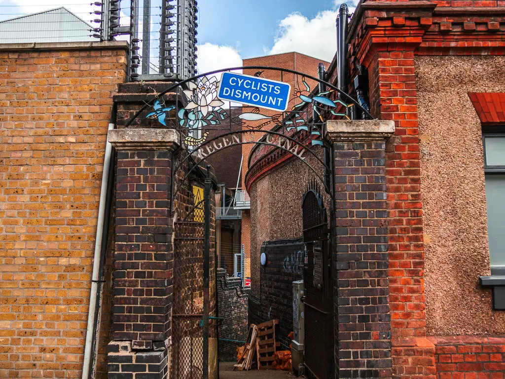 A gap between the brick buildings  with a metal archway entrance to take you down to the regents canal path to continue along the walk. There is a blue sign saying 'cyclists dismount'.