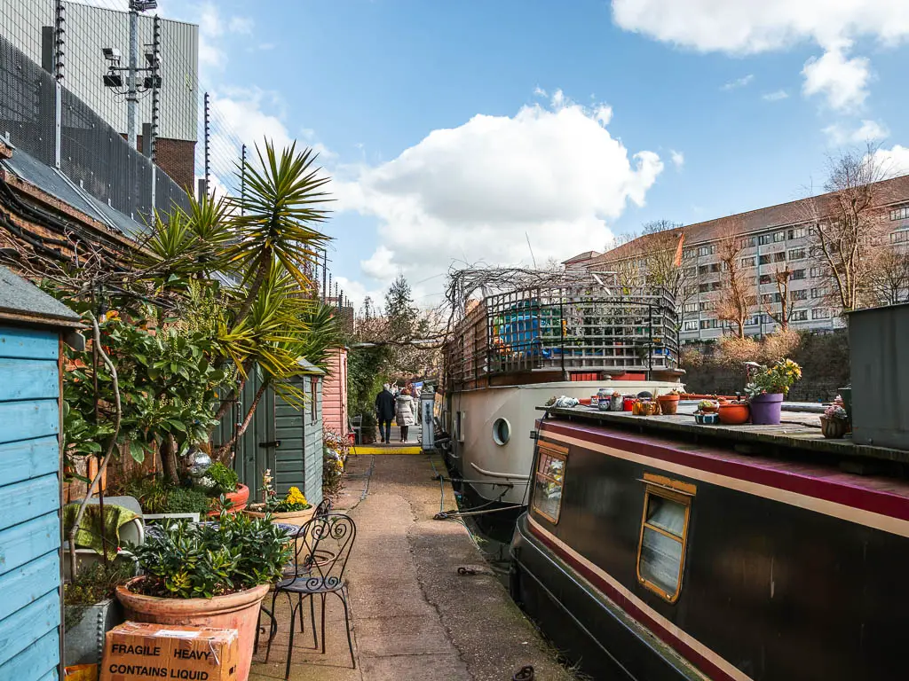 A narrow path lined with barges to the right and mini sheds and plants to the left, along the regents canal walk. There are a couple of people walking ahead on the path. 