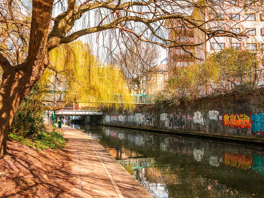 A path along the side of the water, with a graffiti covered brick wall on the other side.