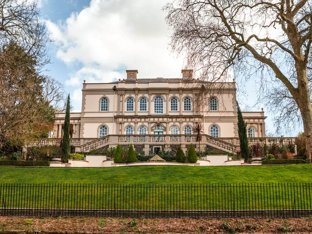 a large mansion with a slopping green grass bank in front, leading to black metal railings.