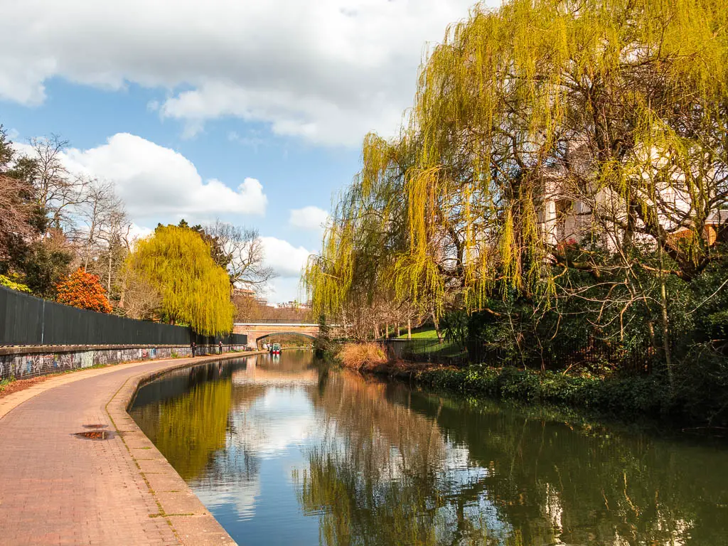 A long stretch of walking path on the left, with the canal on the right. There are overhanging trees on the other side of the canal. There is a bridge visible ahead. 