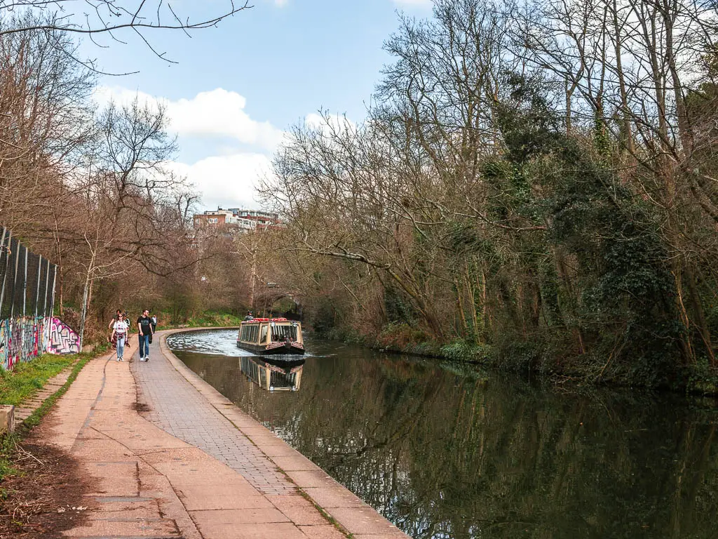 The path on the left of the Regents Canal, with a barge in the middle and a few people walking on the path. The other side of the canal is lined with trees.