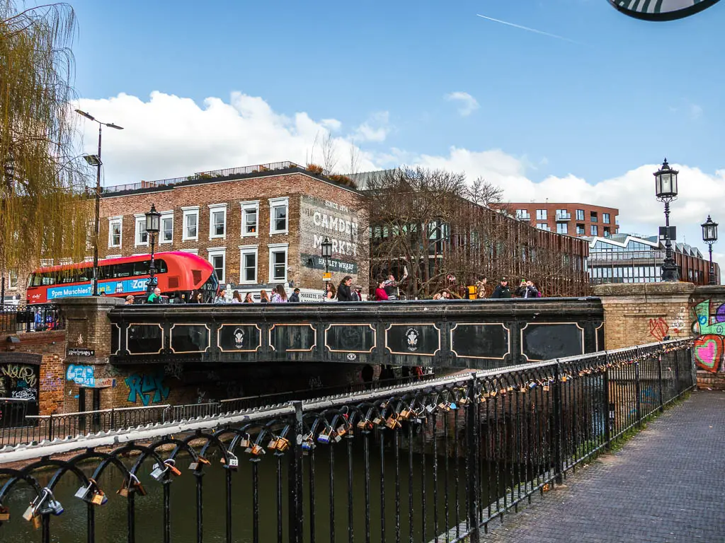 A black metal railing with love locks, and a black bridge ahead. There is a big red bus going over the bridge and lots of people.