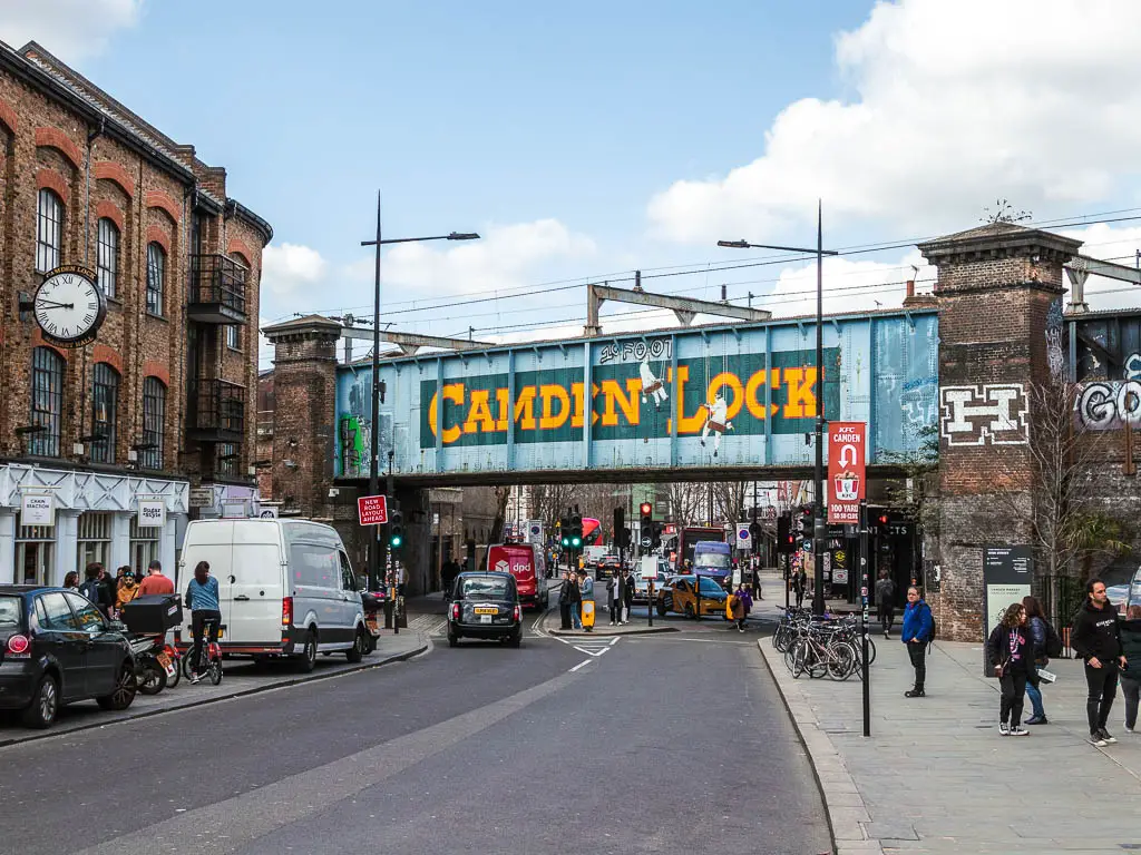 Looking along the road to the big blue railway bridge with Camden Lock written in big yellow letters. There are lots of people walking about.