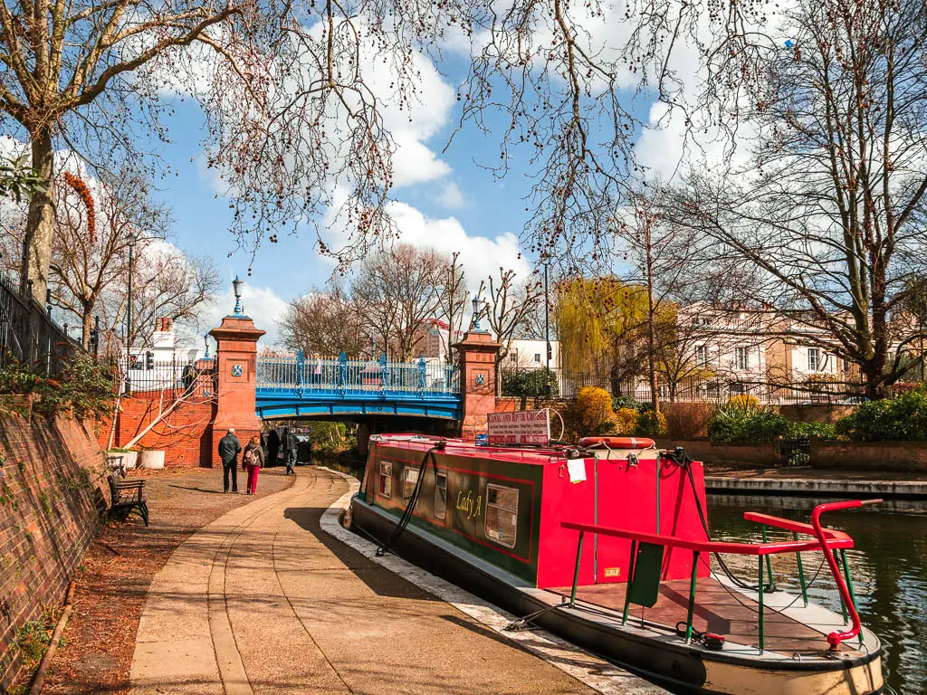 A winding path running under a blue bridge, at the start of the Regents Canal walk. There is a red barge moored to the side of the path.