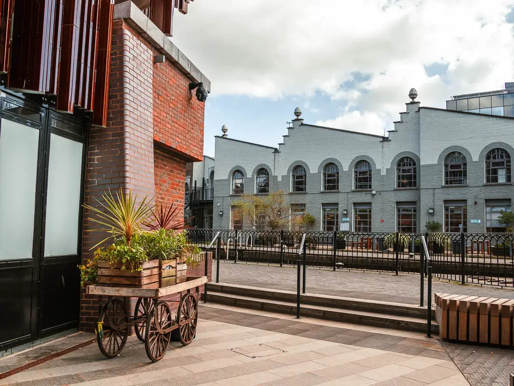 The path with a building on the left and a small wooden trolley with plants in box crates on top. 