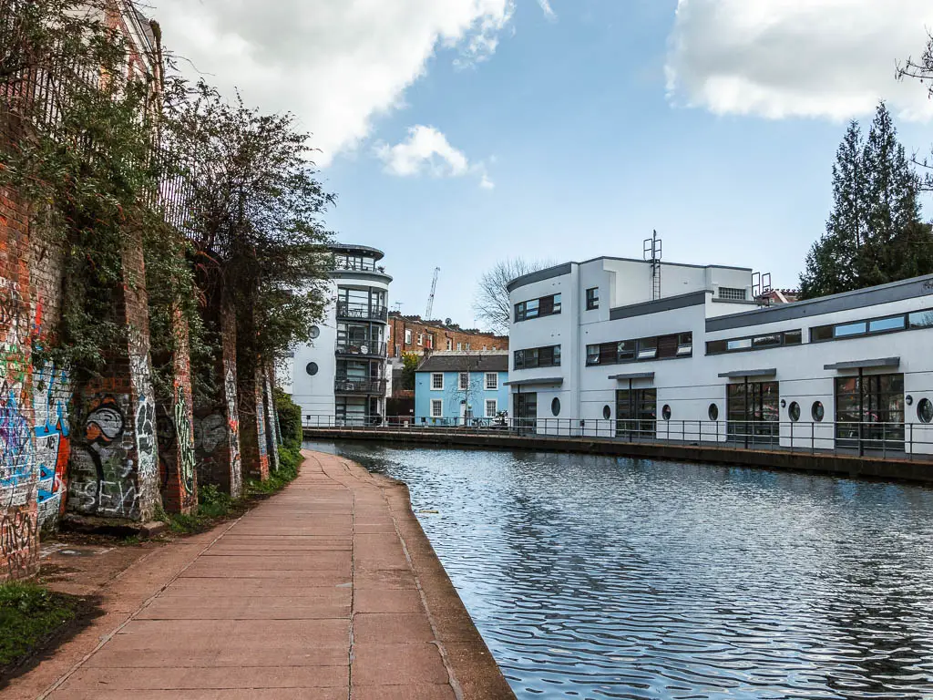 Walking along the regents canal path, with a white coloured building on the other side of the canal, and a small pale blue house next to it.