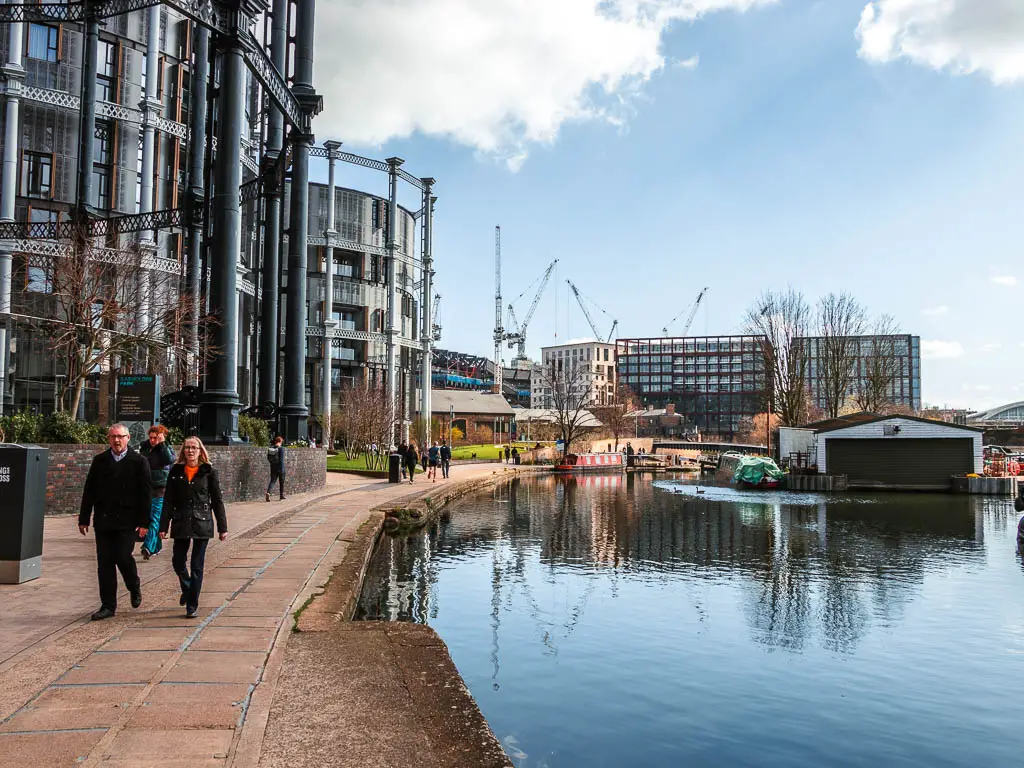 St Pancras basin on the right and the path on the left, with the big metal gass holders and apartments next to the path. There are people walking on the path.